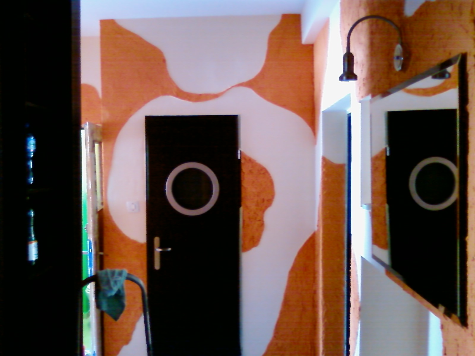 Wnętrze z pomarańczowo-białymi wzorami na ścianie, czarne drzwi z okrągłym oknem i lustro odbijające drzwi, oświetlenie sufitowe i fragment regału z butelkami.